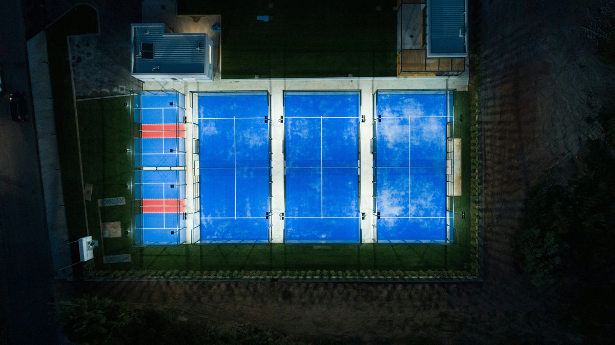 Luftbild einer Padel-Anlage mit beleuchteten blauen Courts bei Nacht